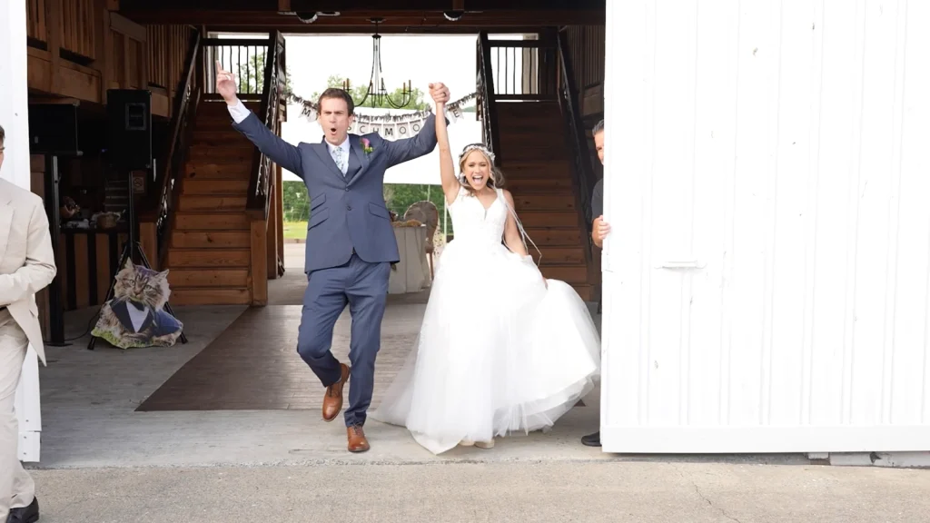 couple enters their reception through a white barn door, cheering with arms raised, at their Rivercest Farm Wedding