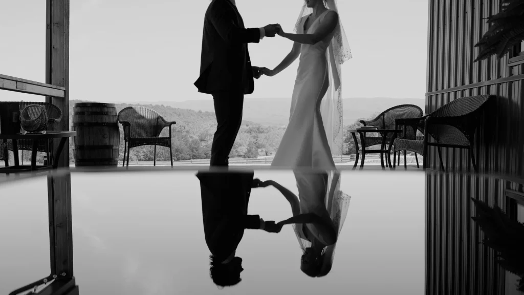 a bride and groom holding hands and dancing, with their reflection in the glass of a table